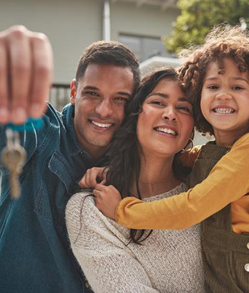 Happy family of three standing outside their new home, smiling and embracing each other while the father holds up a set of house keys.